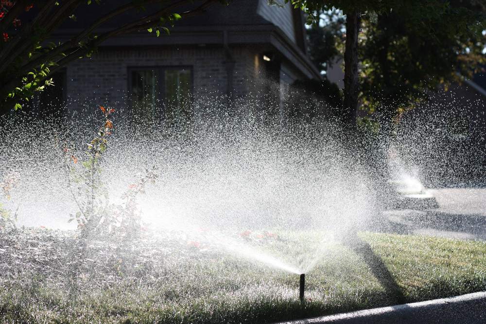 Sprinkler head throwing a wide fan of backlit spray across a lawn